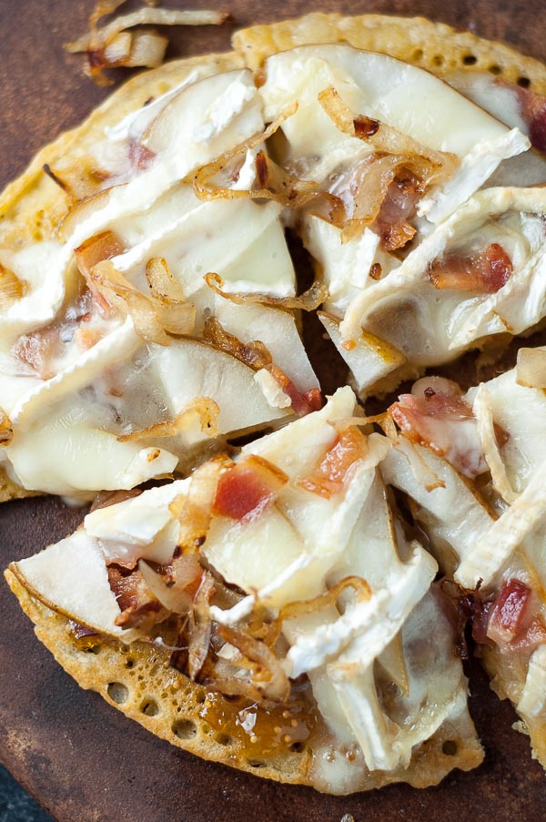 an overhead shot of socca pizza with bacon, brie, and pear on top