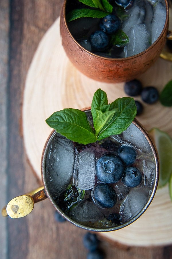 another angle of a overhead shot of two flavored moscow mules on a wooden circle board with blueberries and fresh mint
