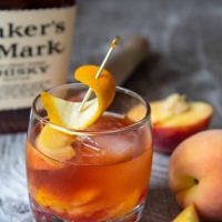 a peach old fashioned on a wood board with bourbon in the background
