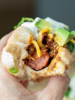 a hand holding up a mexican style hot dog with a bite taken out