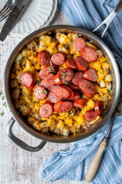 overhead shot of sausage and potatoes in a silver skillet with a blue towel laying next to it