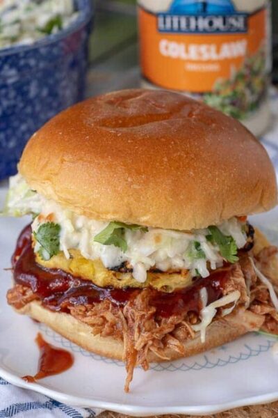 a BBQ pulled chicken sandwich on a plate with coleslaw, BBQ sauce, and grilled pineapple