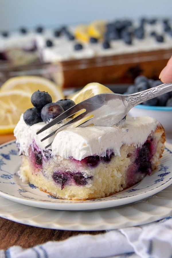 a fork cutting into a piece of lemon blueberry cake on a white plate with fresh blueberries on top
