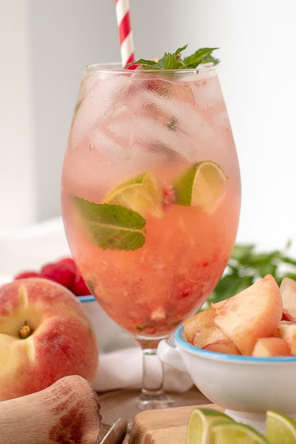 A Raspberry Peach Mojito in a tall glass with cut up peaches and muddler next to it