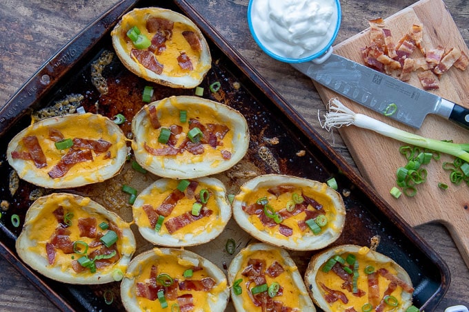 an overhead shot of loaded potato skins on a baking sheet with sour cream and green onions next to it