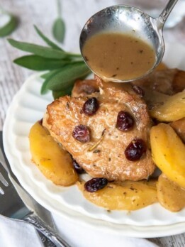 pork medallions on a white plate with apples and cranberries and a ladle of dijon gravy being poured over