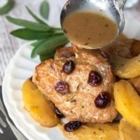 pork medallions on a white plate with apples and cranberries and a ladle of dijon gravy being poured over