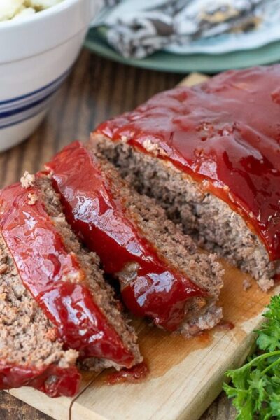 instant pot meatloaf sliced on a wooden board with a bowl of mashed potatoes in the background