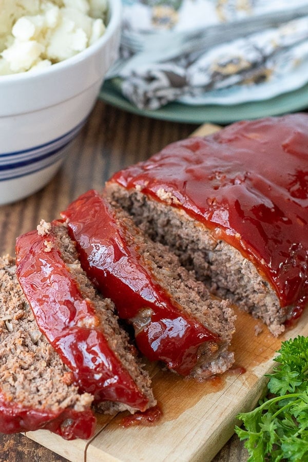 meatloaf sliced on a wooden board with a bowl of mashed potatoes in the background