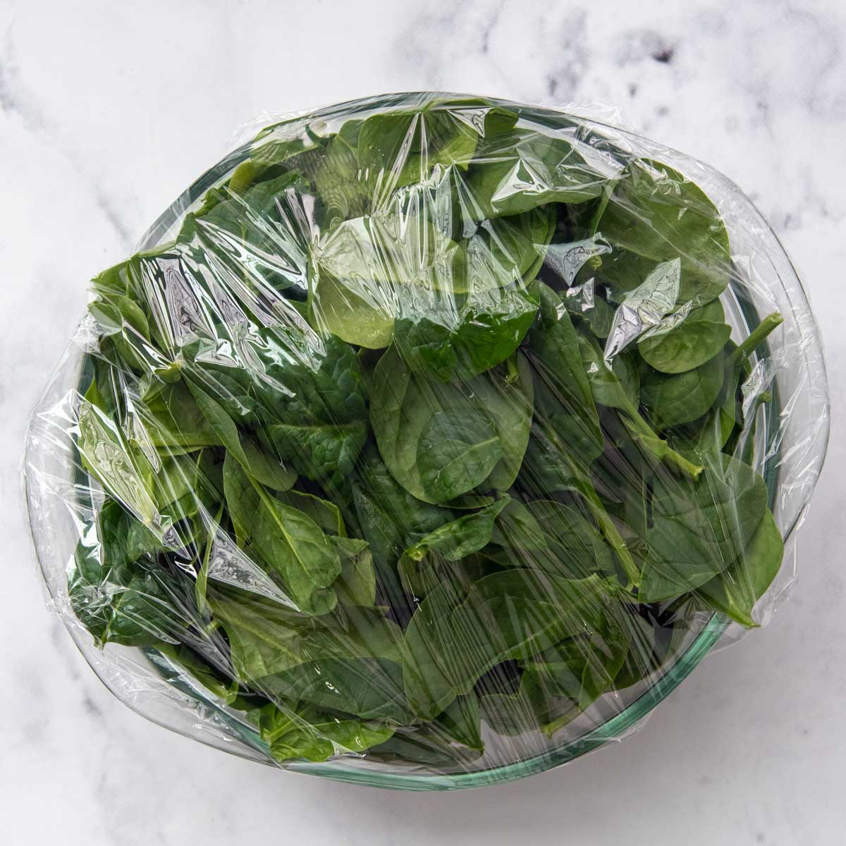 fresh spinach in a glass bowl covered with plastic wrap