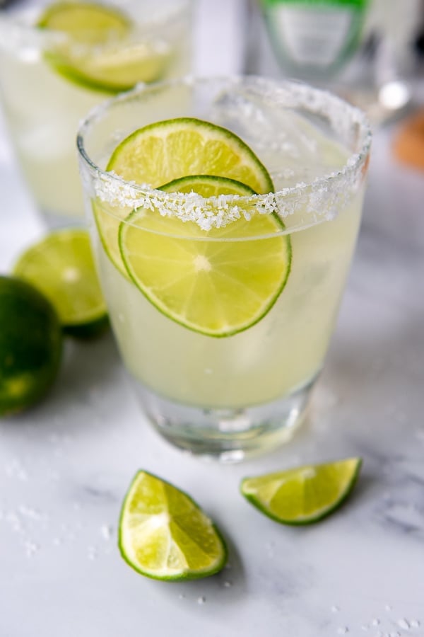a glass of limeade margarita on a white marble surface with cut limes around