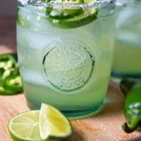 a margarita on a wooden cutting board with fresh jalapenoes and limes around it