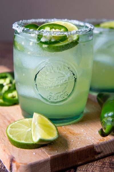 a margarita on a wooden cutting board with fresh jalapenoes and limes around it