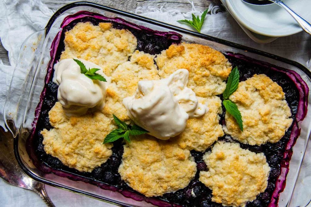 horizontal picture of blueberry cobbler in a glass dish with melting ice cream on top
