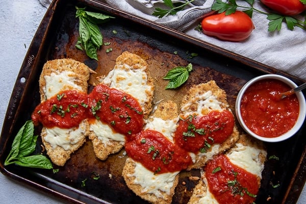 overhead shot of a baking sheet of gluten free chicken parmesan with fresh basil scattered around and a cup of marinara