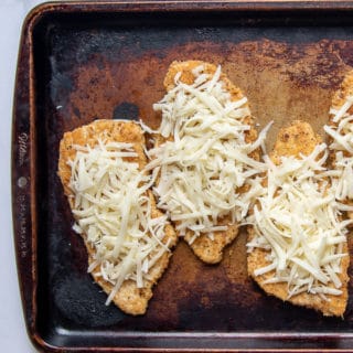 breaded chicken breasts on a baking sheet with grated cheese on top