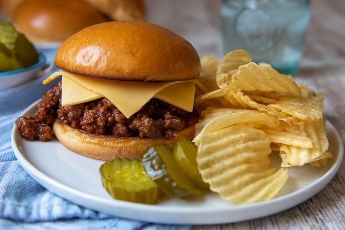 a sloppy joe on a plate with potato chips and pickle slices