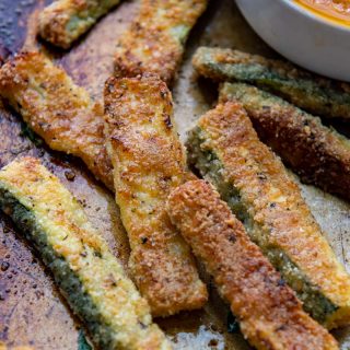 close up of baked zucchini fries on a baking sheet with dip in background