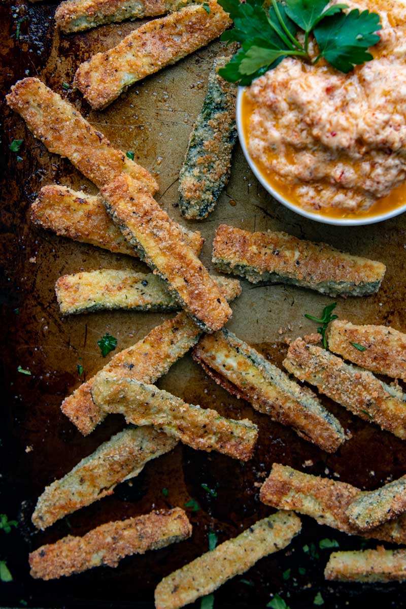 overhead shot of baked zucchini fries on a sheet pan with a small white bowl of dipping sauce