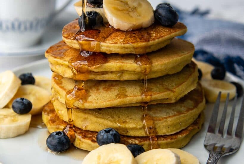 syrup being poured on a stack of pancakes with bananas and blueberries scattered around.