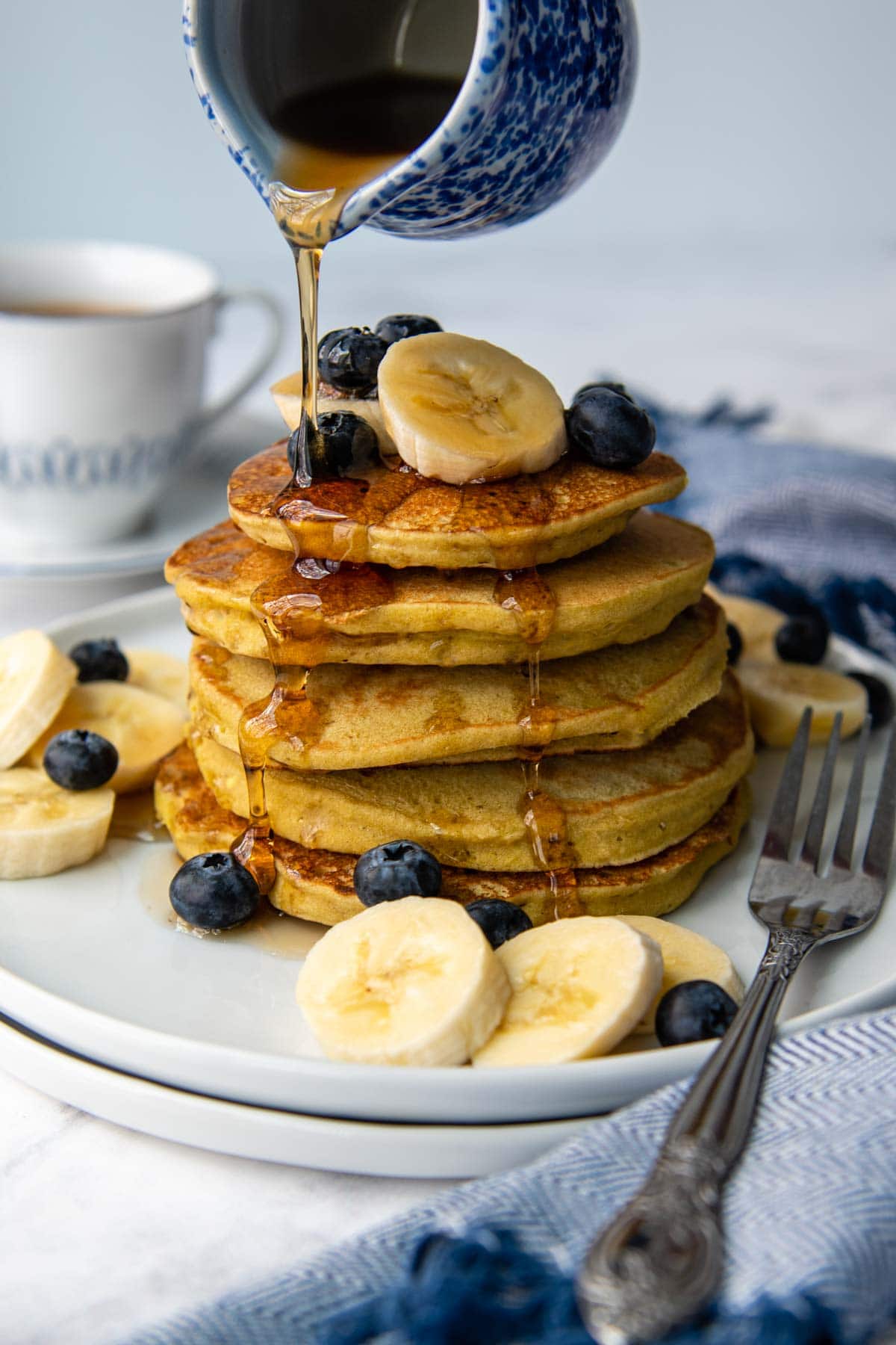 syrup being poured on a stack of pancakes with bananas and blueberries scattered around. 