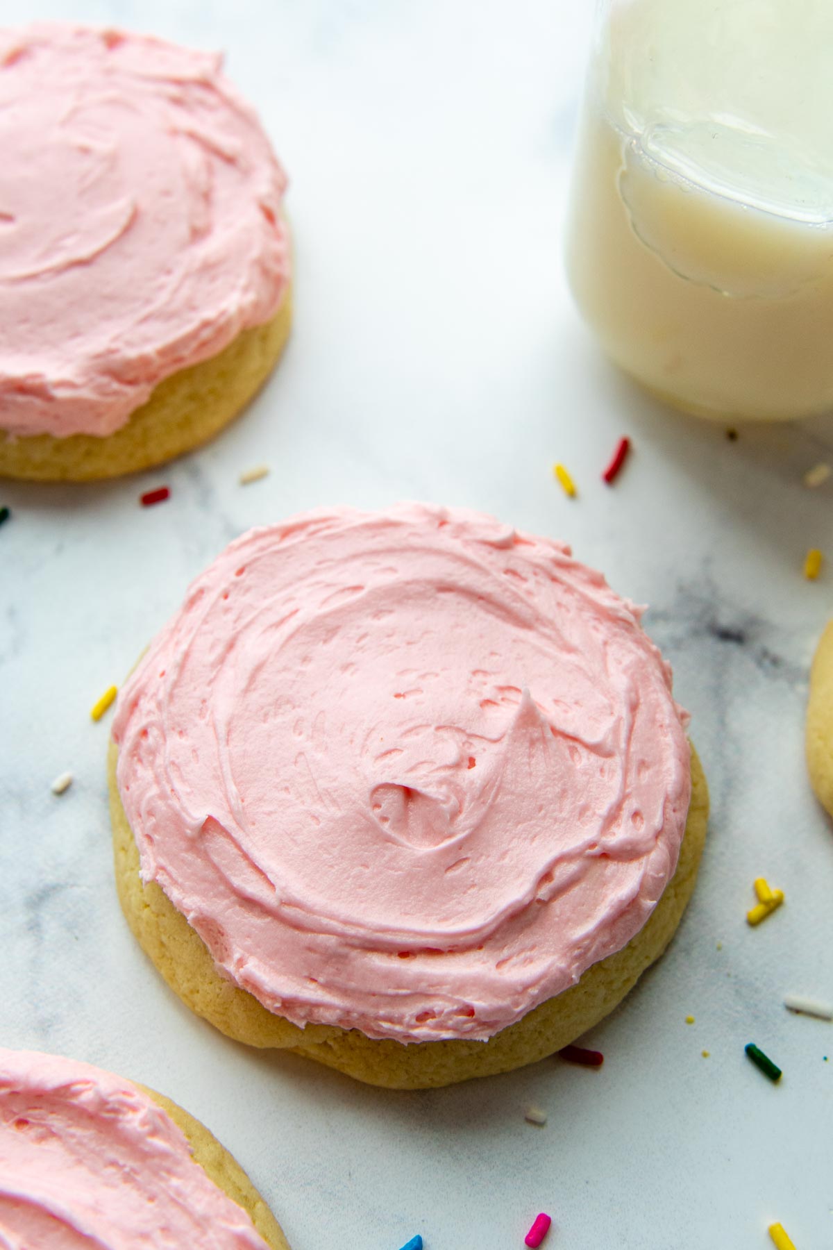 a pink sugar cookie on a white background with sprinkles around and a glass of milk.