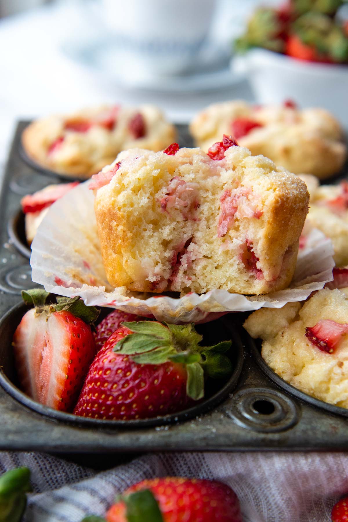 a bitten strawberry muffin resting on a muffin pan with fresh strawberries in one of the cups.