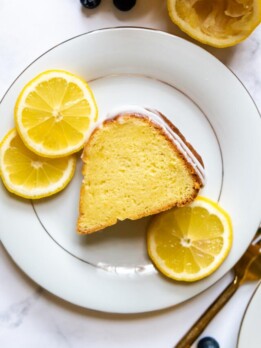 a slice of lemon cake on a white plate with lemon slices next to it.