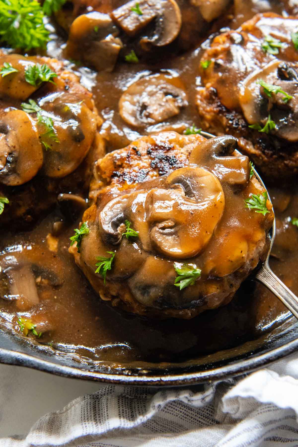 a spoon lifting up a salisbury steak with mushrooms on top from a skillet.