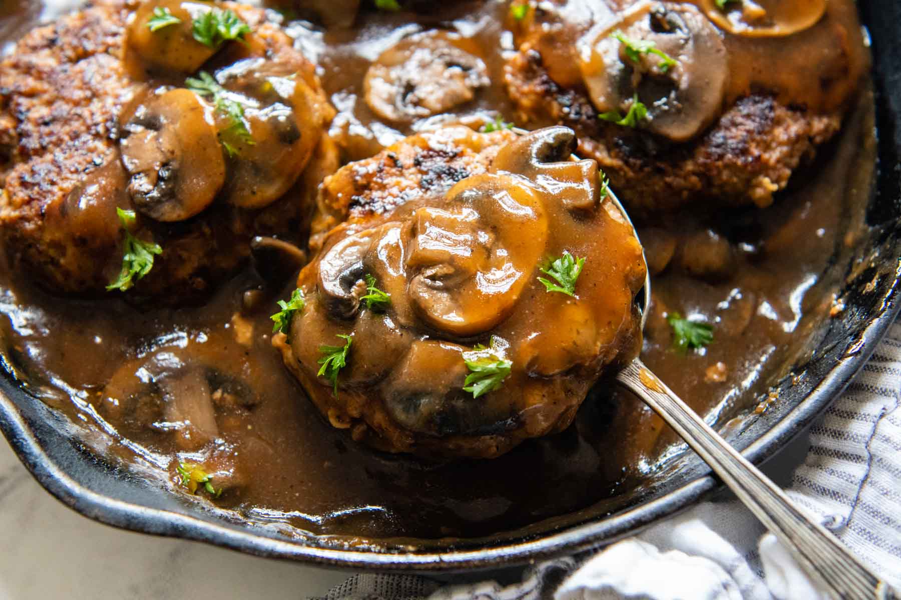 a spoon lifting up a salisbury steak with mushrooms on top from a skillet.