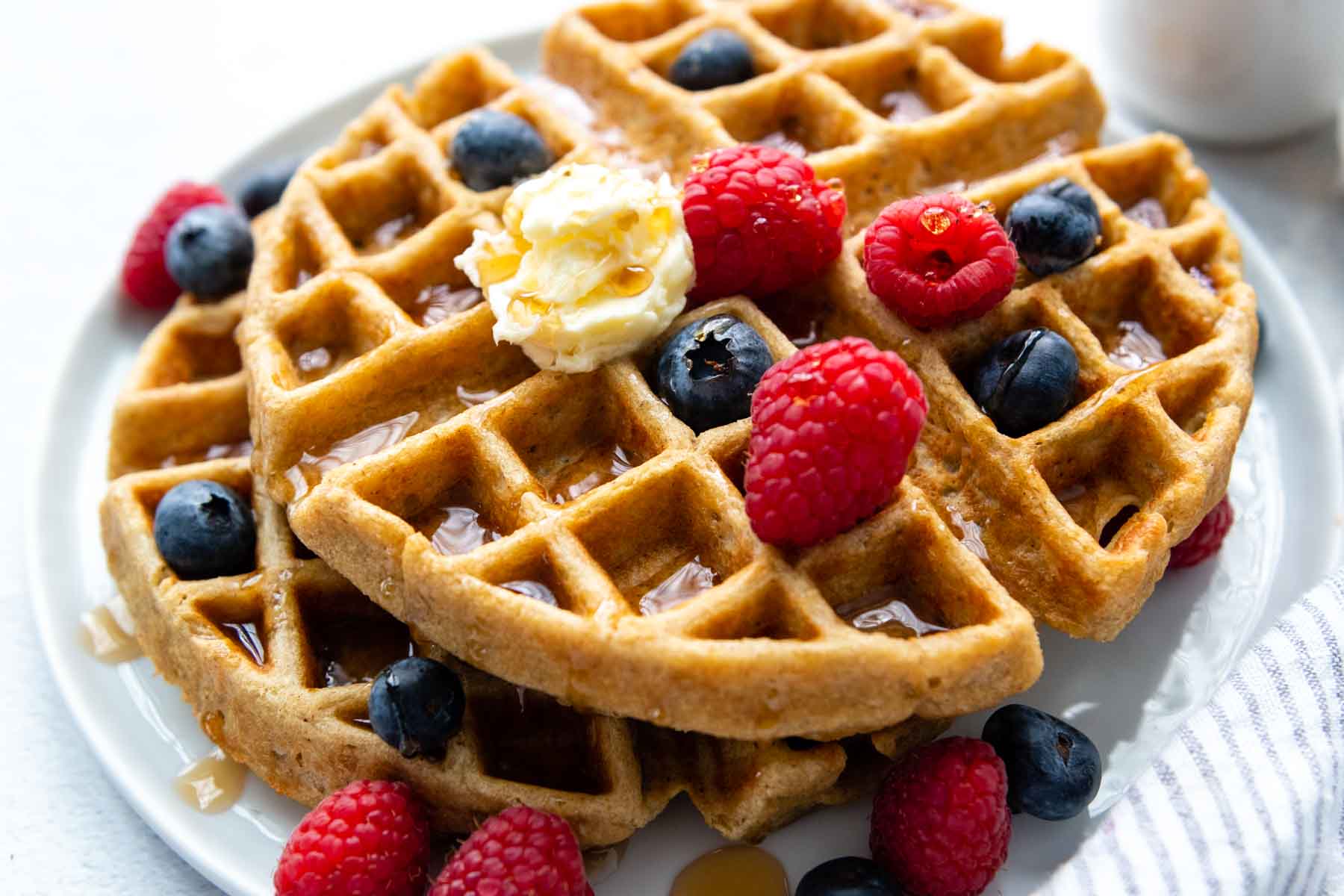 a stack of waffles on a white plate with berries, butter and syrup.