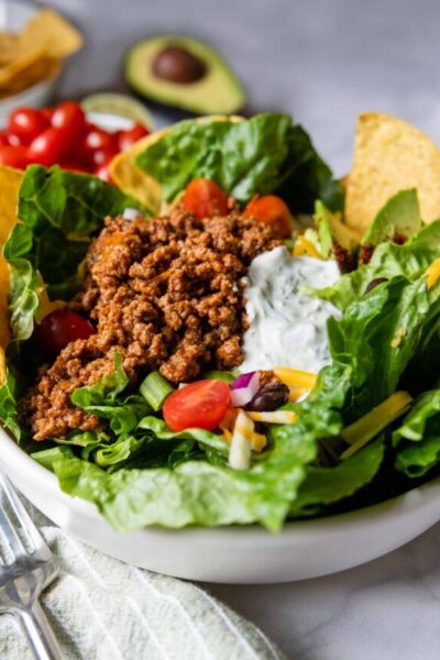 a taco salad in a white bowl with fixings in the background.