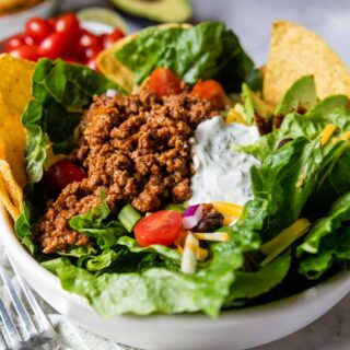 a taco salad in a white bowl with fixings in the background.