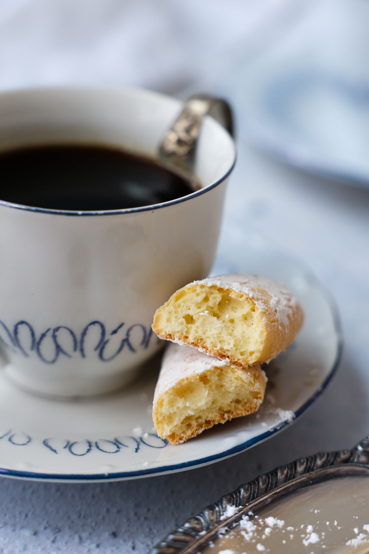 A lady finger broke in 1/2 showing the inside texture while sitting on a saucer next to a cup of tea.