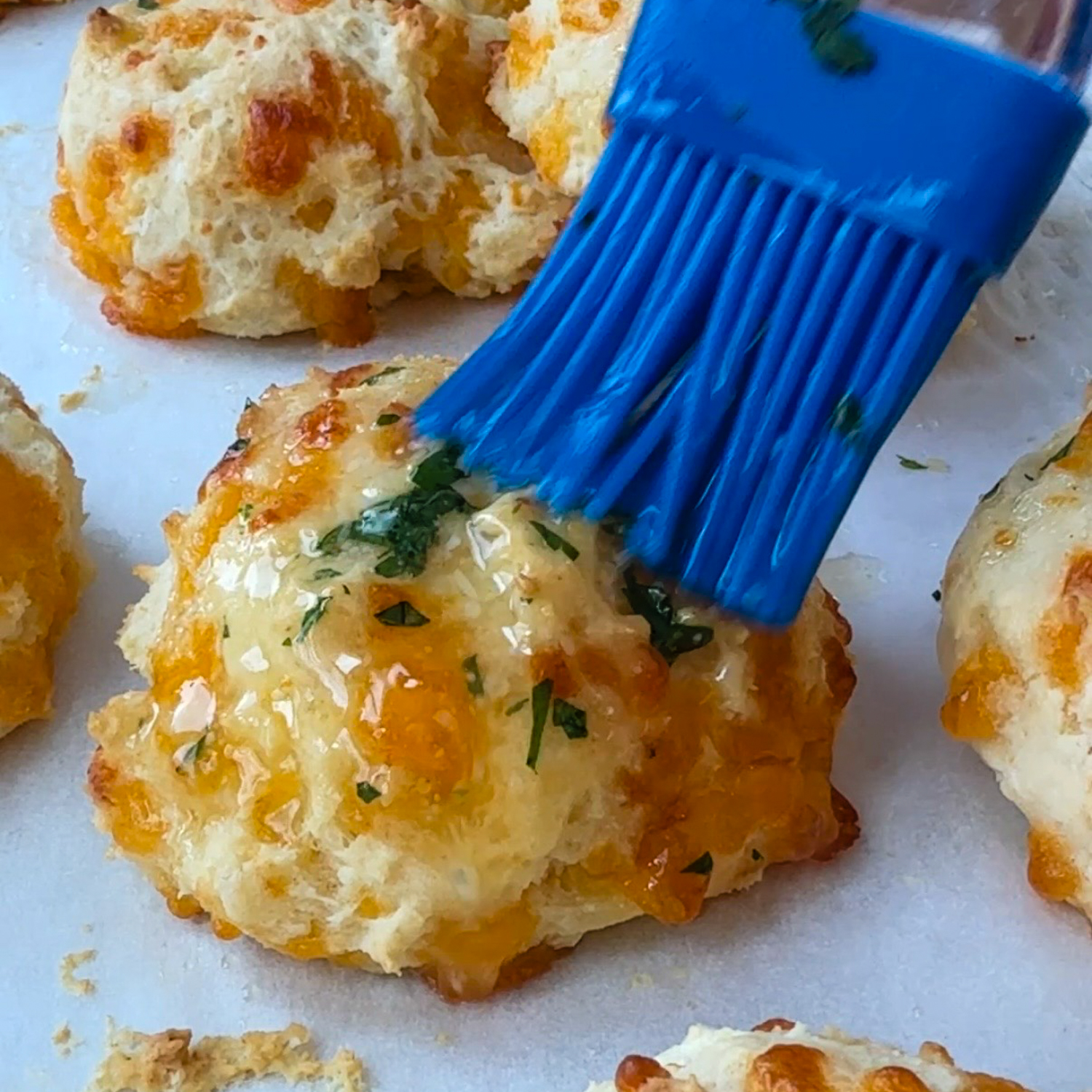 Blue basting brush spreading butter on cooked biscuits.
