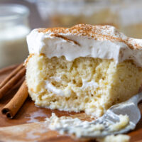 An slice of tres leches cake on a wooden plate with a bite removed showing the texture of the inside of the cake.