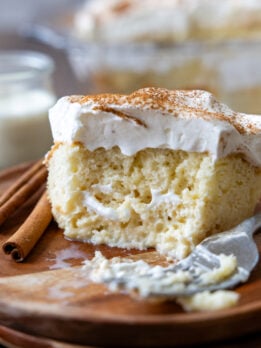 An slice of tres leches cake on a wooden plate with a bite removed showing the texture of the inside of the cake.