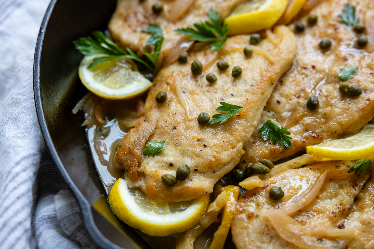 Chicken cutlet in a skillet with lemon garnish and topped with capers.