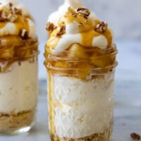 Mini cheesecake in a glass jar with apples, a dollop of whipped cream, caramel, and pecans on top, with pecans scattered around it.