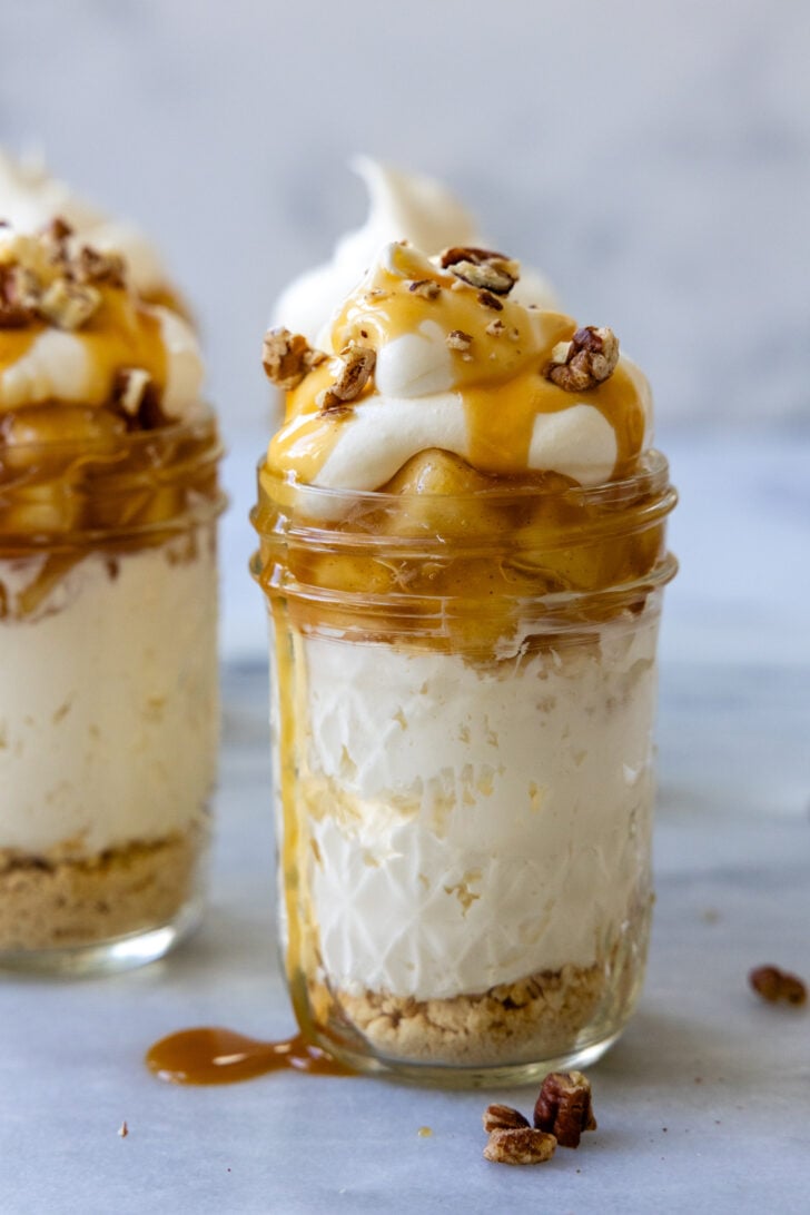 Mini cheesecake in a glass jar with apples, a dollop of whipped cream, caramel, and pecans on top, with pecans scattered around it.