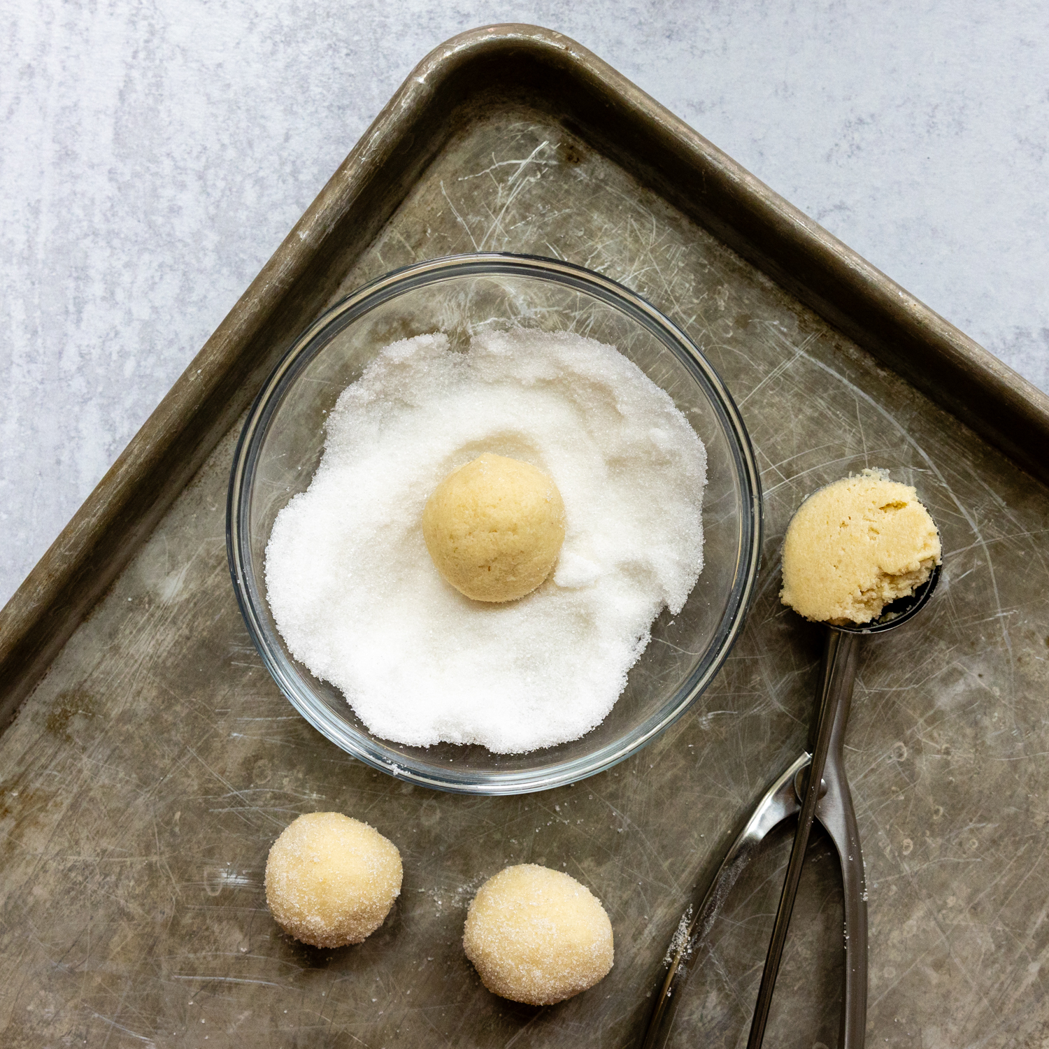 Rolling dough in a glass bowl filled with sugar on a baking sheet with a cookie scoop.